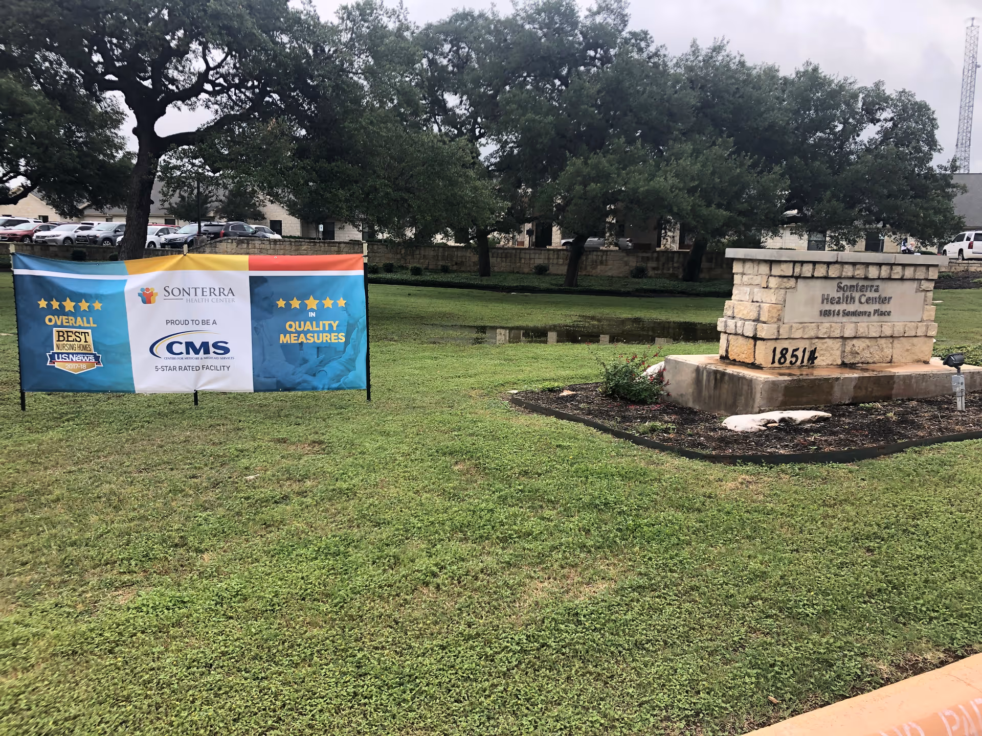 Outdoor view of Sonterra Health Center showing a stone sign with the facility name and address 18514 Sonterra Place, next to a banner highlighting the center's 5-star rating by CMS and recognition for overall best nursing homes and quality measures. The area has green grass, trees, and parked cars in the background.