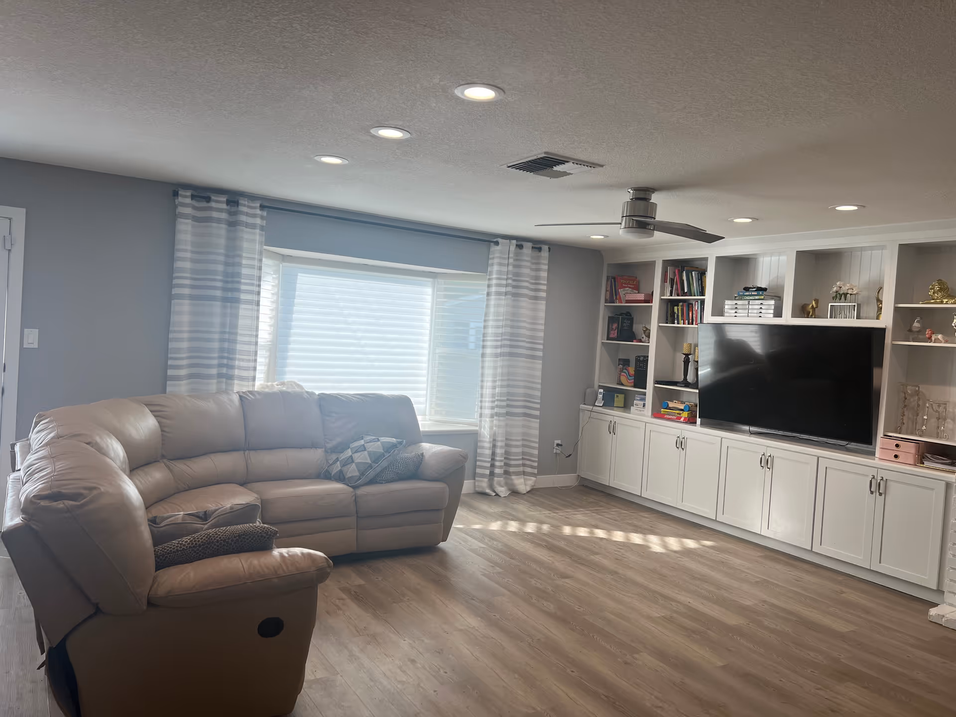 Bright living room with a beige leather sectional, large window with striped curtains, built-in white shelving and a TV over wood-look flooring.