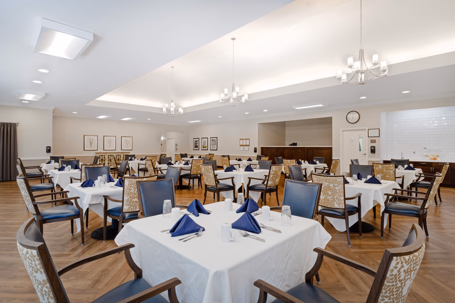 A spacious senior living dining room with multiple tables covered in white tablecloths, each set with blue folded napkins, glasses, and silverware. The room features wooden chairs with patterned backs and blue seats, wooden flooring, and modern ceiling lights. The walls are decorated with framed artwork and a clock is visible above a door.