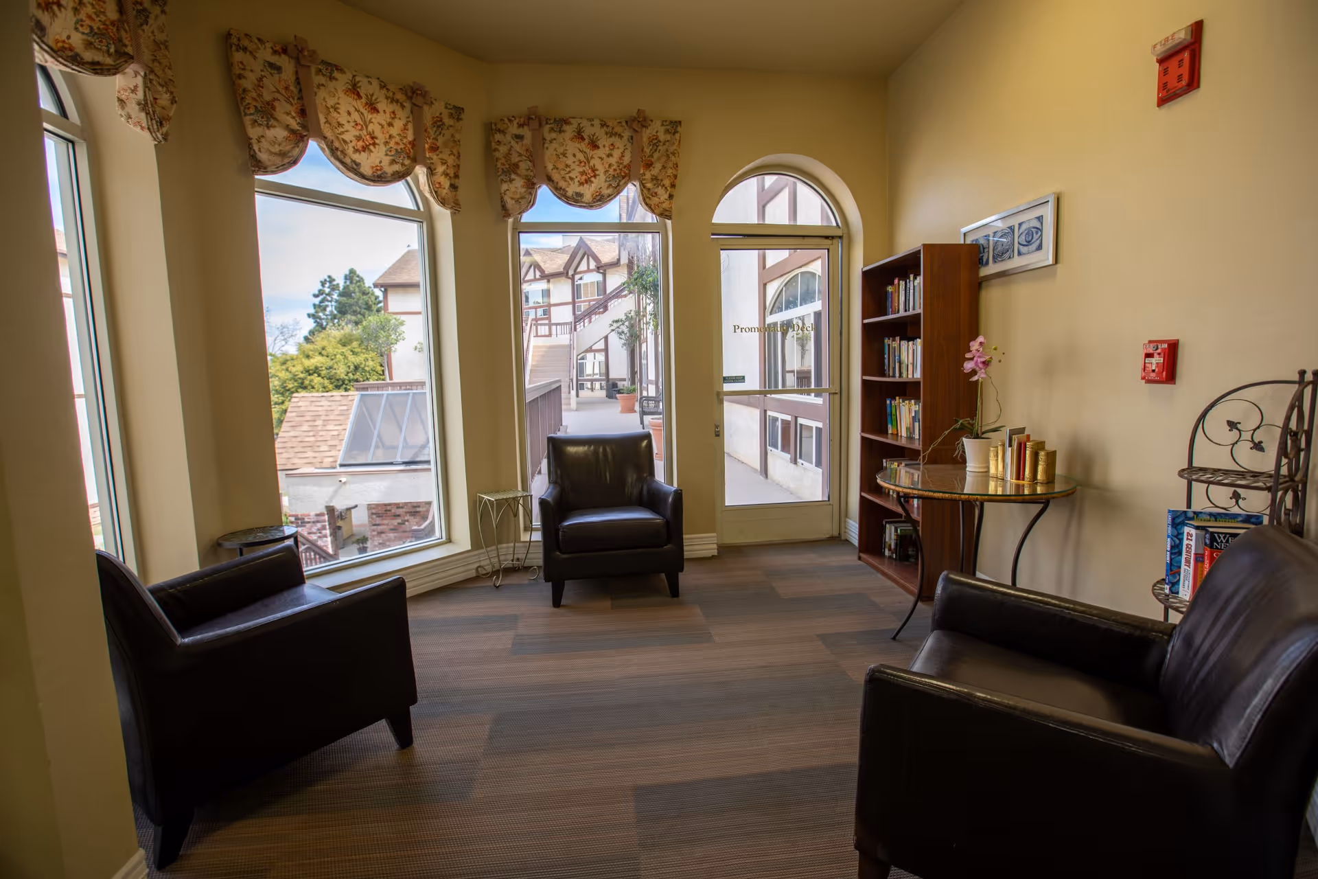 A cozy sitting area with three black leather armchairs arranged around a small glass table with books and a potted orchid. The room has large windows with floral valances letting in natural light and a door leading to an outdoor walkway. A wooden bookshelf filled with books is against the wall.