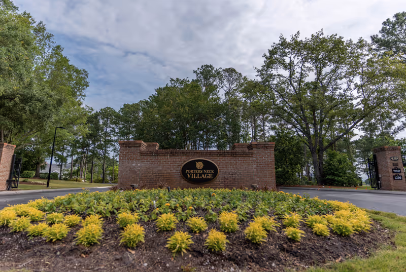 Entrance to Porters Neck Village with a brick sign surrounded by landscaped plants and trees under a partly cloudy sky.