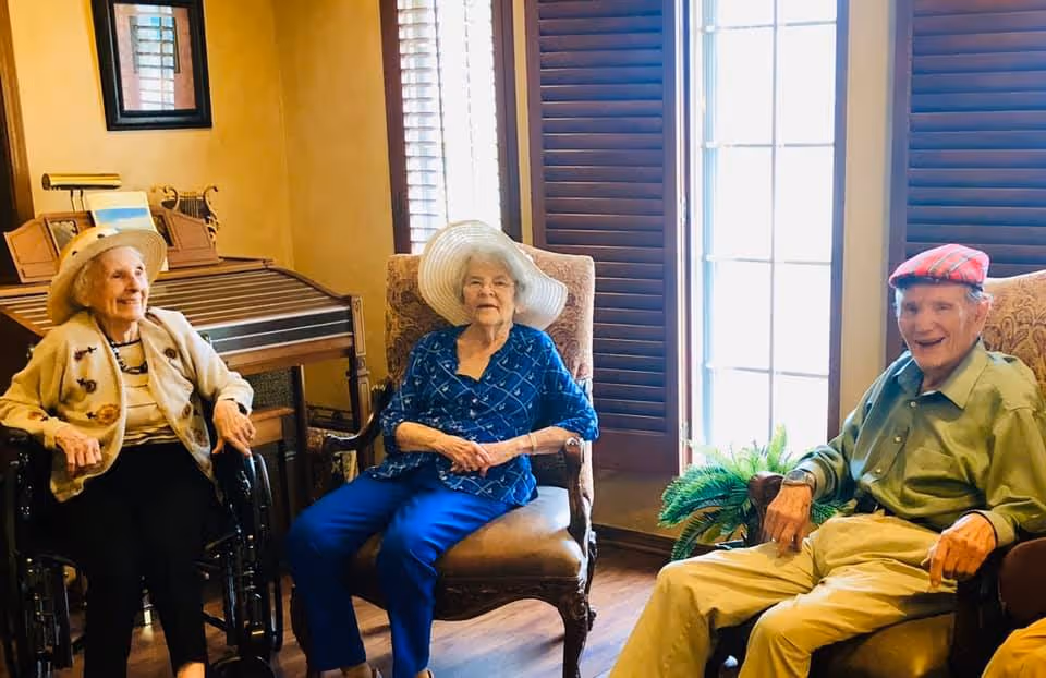 Three elderly individuals sitting indoors near large windows with wooden shutters. Two women are seated in chairs, one wearing a white wide-brimmed hat and blue outfit, the other in a wheelchair wearing a beige jacket and hat. A man wearing a red plaid cap and green shirt is seated in a chair. There is a small plant near the window and a wooden piece of furniture with framed pictures in the background.