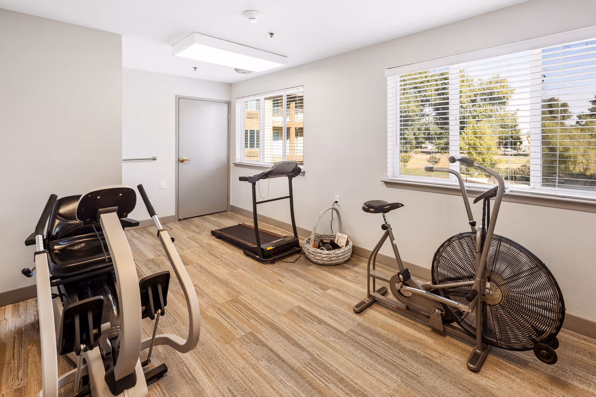 A small exercise room with a recumbent exercise bike, a treadmill, and a stationary fan bike. The room has beige walls, a large window with white blinds showing trees outside, and a door with a silver handle. The floor is covered with light brown patterned carpet.