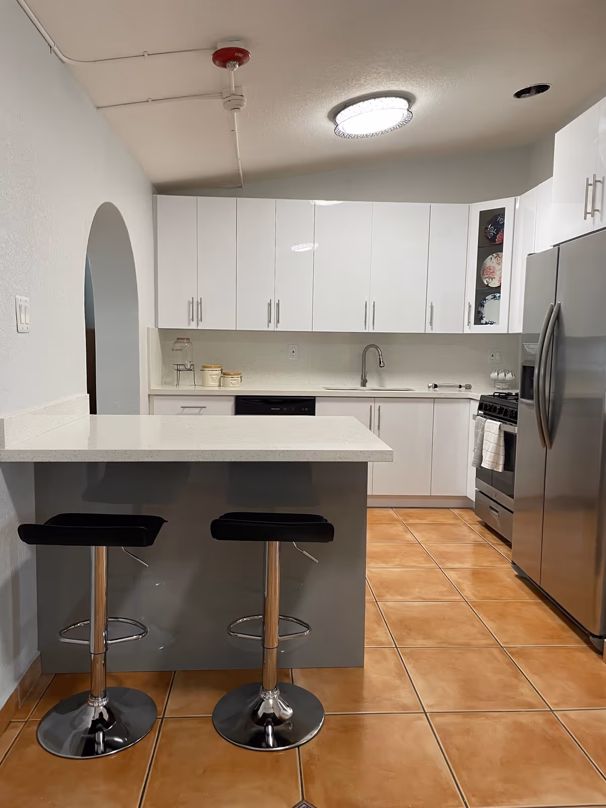 Modern kitchen with white cabinets, a stainless steel refrigerator, a stove, and a kitchen island with two black bar stools. The floor is tiled with brown tiles and there is a round ceiling light fixture.