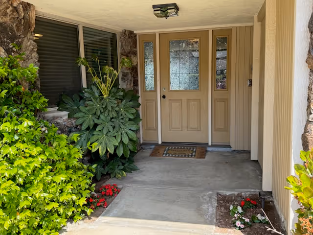 Front entrance of a building with a beige door featuring glass panels, surrounded by stone and beige siding. There are green leafy plants and small flower beds on either side of the concrete walkway leading to the door.