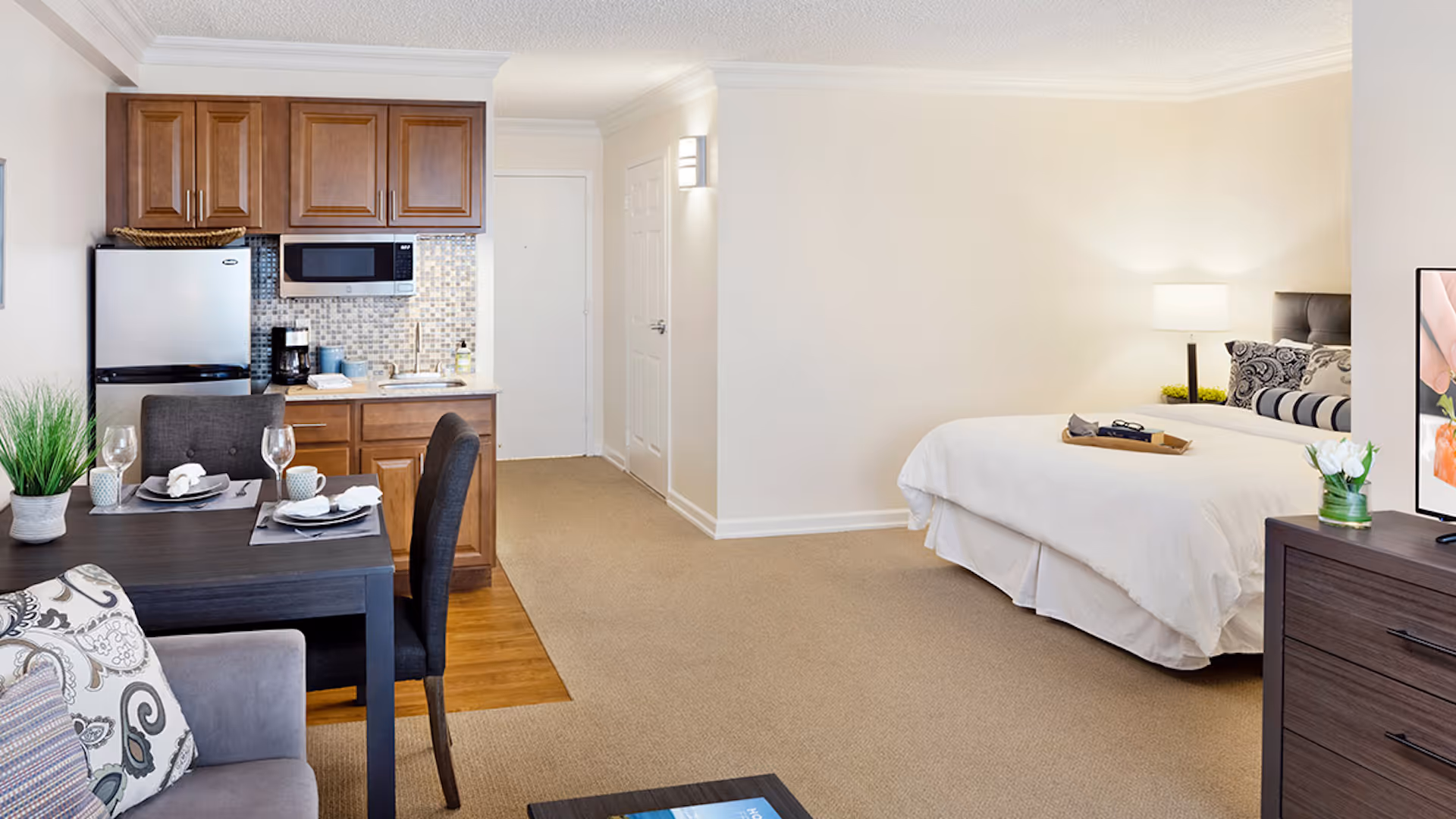 A studio apartment interior featuring a small kitchen with wooden cabinets, a refrigerator, microwave, and coffee maker. Adjacent to the kitchen is a dining table set for two with wine glasses and plates. The living area includes a sofa with patterned cushions, and a bedroom area with a neatly made bed, bedside lamp, and a TV on a dresser. The room has beige walls and carpeted flooring.