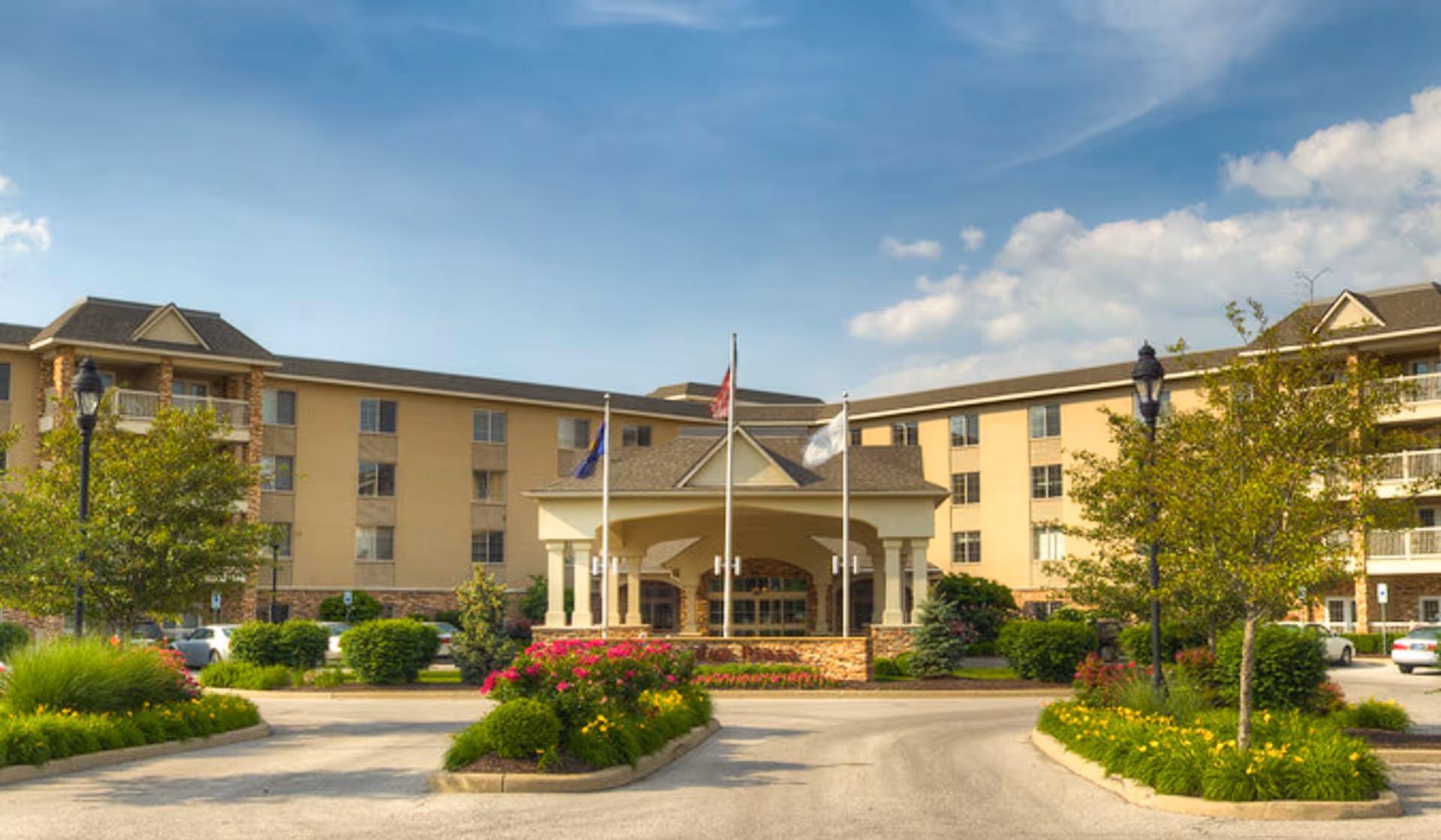 Front exterior view of Westside Garden Plaza, a multi-story senior living facility with a covered entrance, three flagpoles, landscaped flower beds, trees, and a clear blue sky.