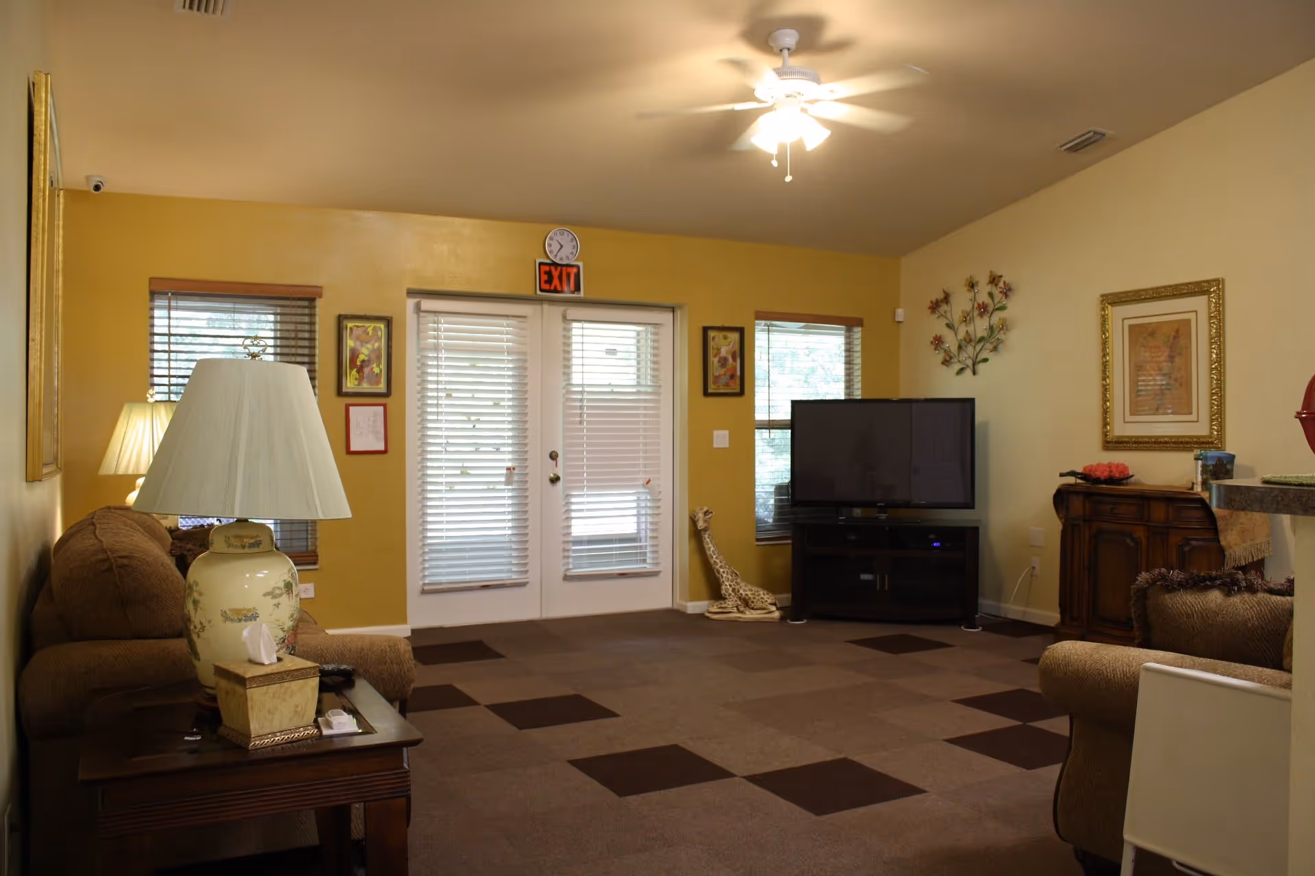 A cozy living room with brown sofas, a side table with a lamp and tissue box, a flat-screen TV on a stand, decorative wall art, and double doors with blinds leading outside. The room has a ceiling fan with a light and windows with wooden blinds.