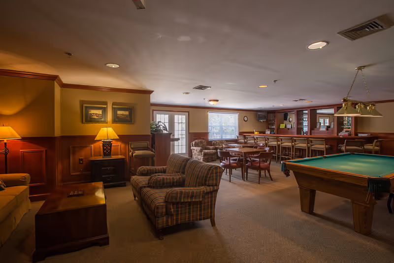 A cozy common area with a pool table, several armchairs, a coffee table, a bar with stools, and a few small round tables with chairs. The room has warm lighting, wood paneling on the walls, and windows letting in natural light.