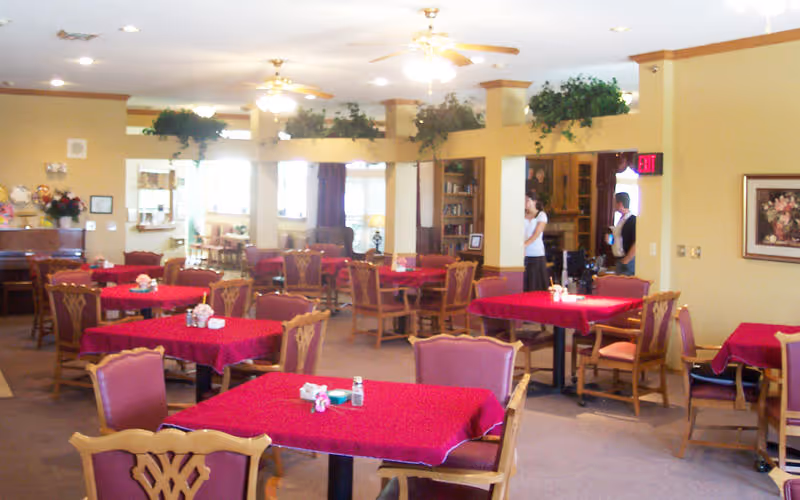 A spacious dining room with multiple tables covered in red tablecloths, surrounded by wooden chairs with cushioned seats. The room has beige walls, ceiling fans with lights, and some greenery on top of a partial wall divider. Two people are visible in the background near a doorway.