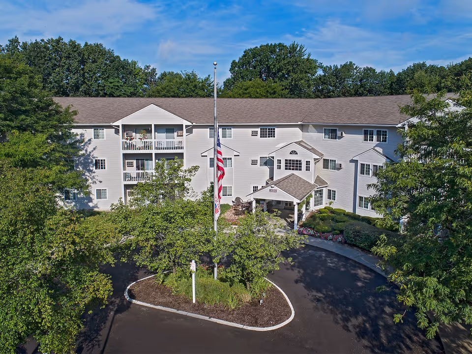 Exterior view of a three-story senior living facility building surrounded by trees and greenery. The building has a light-colored facade with multiple windows and balconies. In front of the building is a circular driveway with a flagpole displaying the American flag. The sky is clear with some clouds.