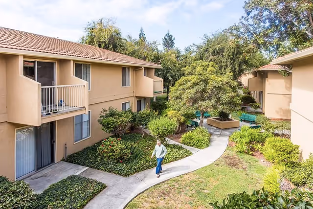 Outdoor courtyard area of a retirement community with beige two-story buildings, green shrubs, trees, and a paved walkway. A person is walking on the path, and there are green benches near a tree in the center.