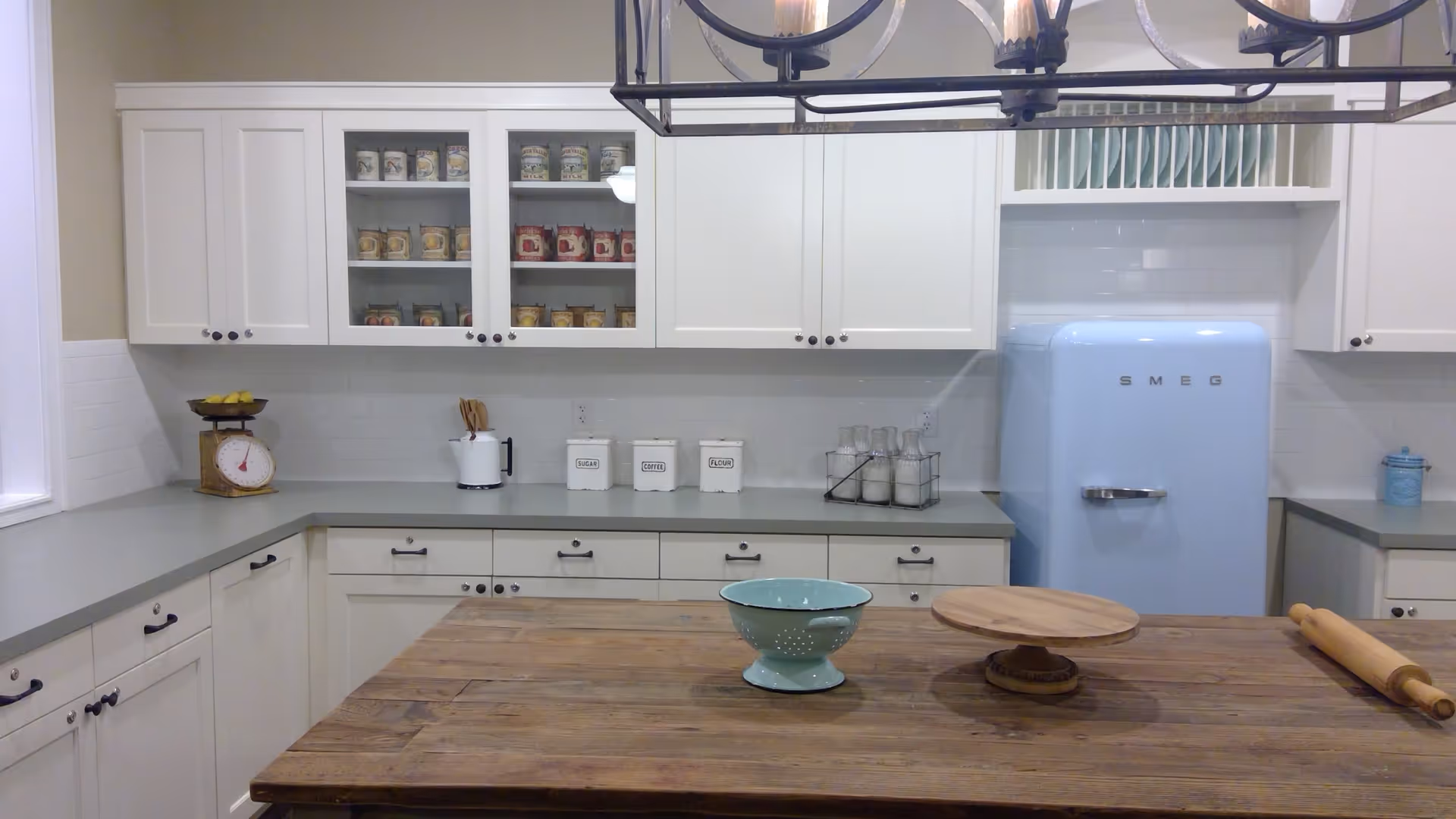 A modern kitchen with white cabinets, a light blue SMEG refrigerator, a wooden island countertop with a rolling pin, a wooden cake stand, and a light blue colander. The backsplash is white subway tile, and there are various containers and kitchen utensils on the counters.