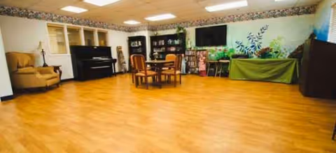 Spacious senior living activity room with chairs, piano, bookshelves, and plants.