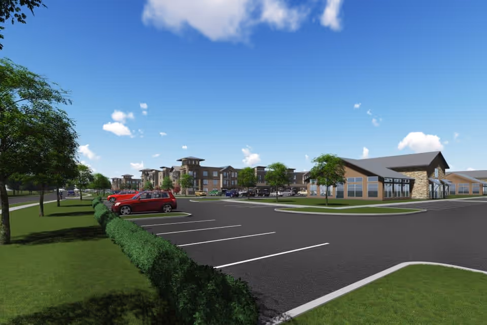 View of a senior living facility parking lot with several parked cars, landscaped greenery including trees and bushes, and multi-story residential buildings in the background under a blue sky with scattered clouds.