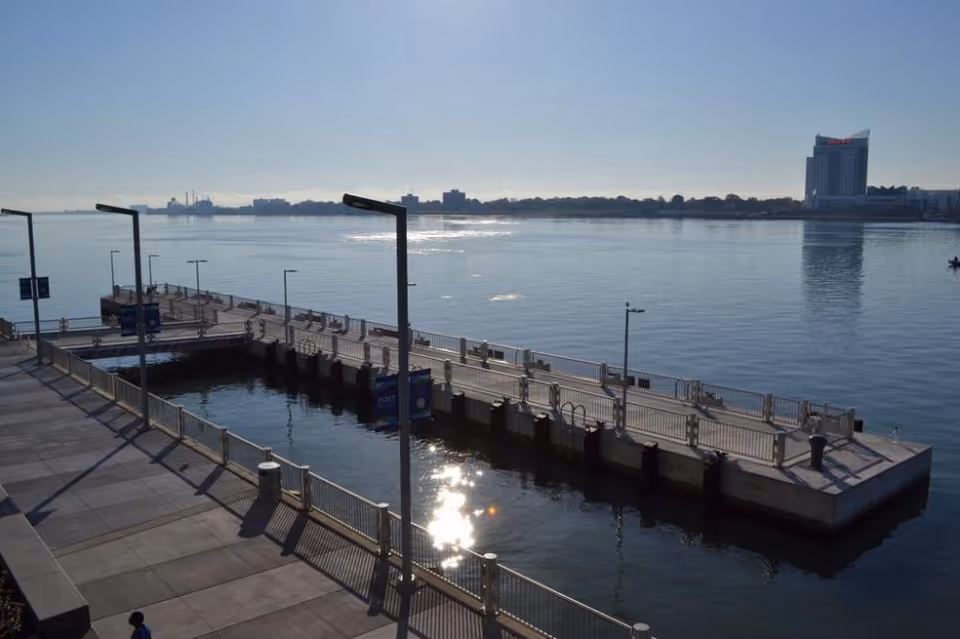 Sunlit waterfront pier with railings and lampposts extending into calm water and a distant city skyline.