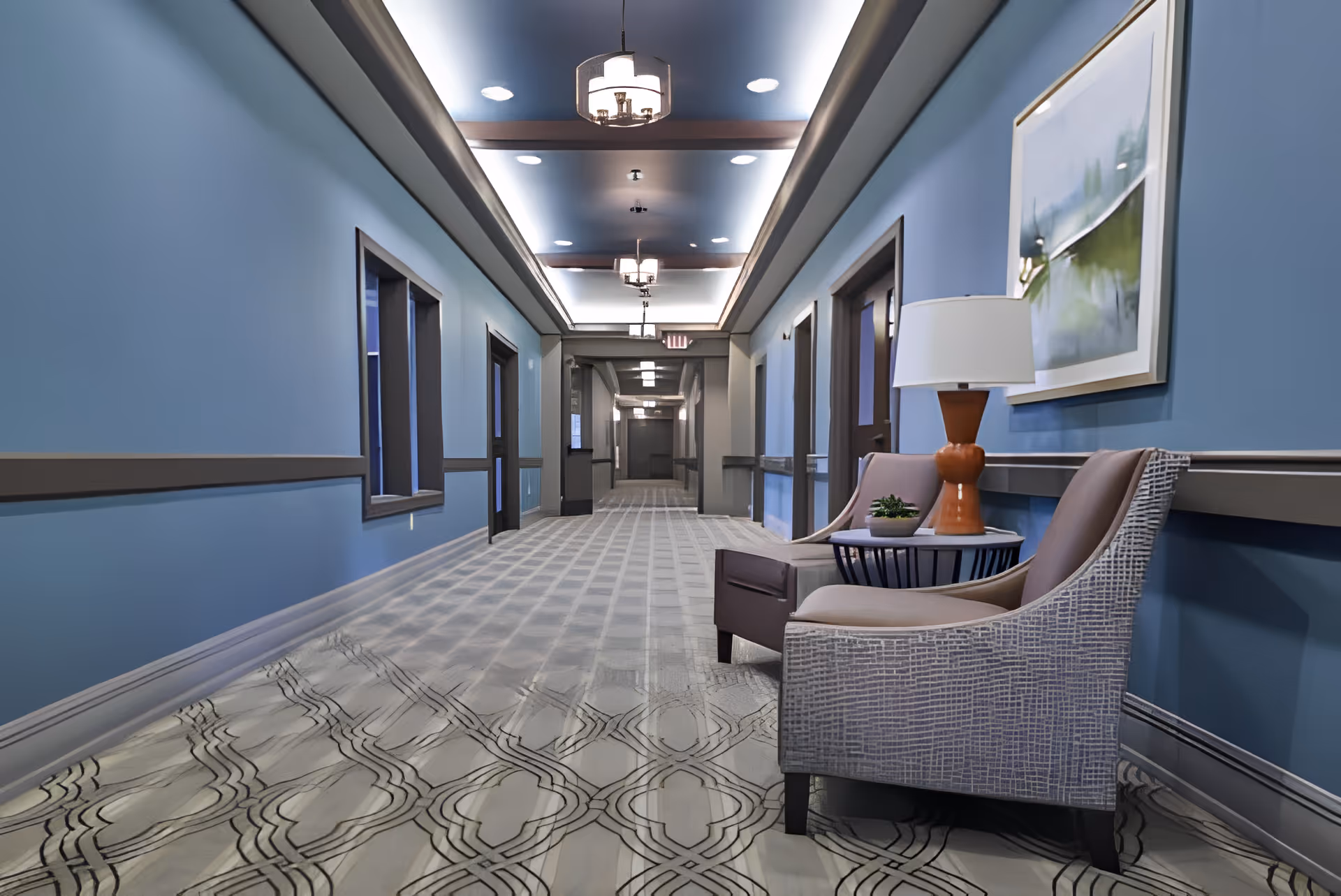 A long, well-lit hallway with blue walls and patterned carpet. On the right side, there are two cushioned chairs with a small round table between them, holding a lamp and a small plant. The ceiling features recessed lighting and decorative light fixtures. Several doors and windows line the hallway.