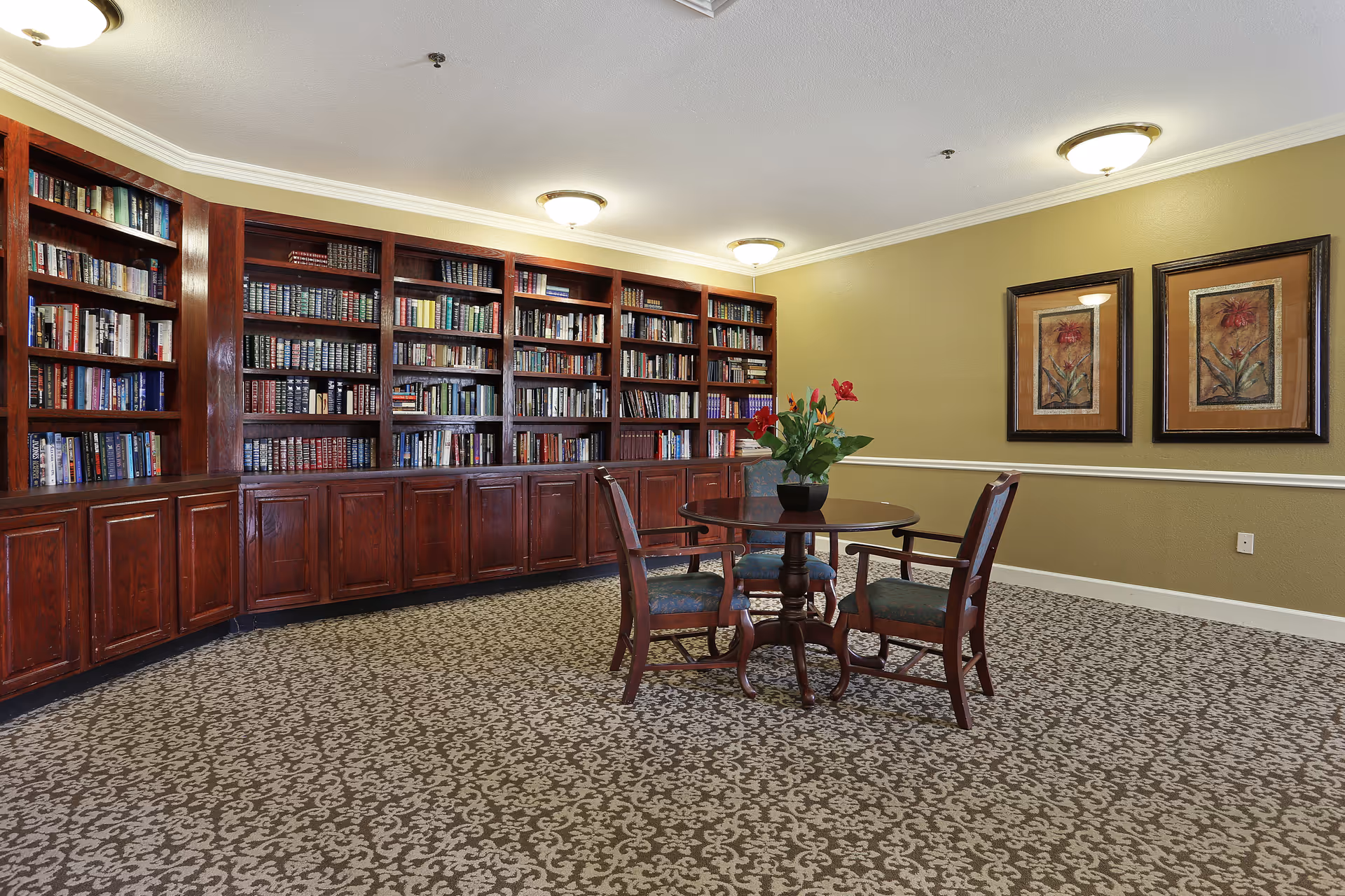 Quiet communal library with built-in wooden bookshelves, a round table with four chairs and framed artwork on the wall.