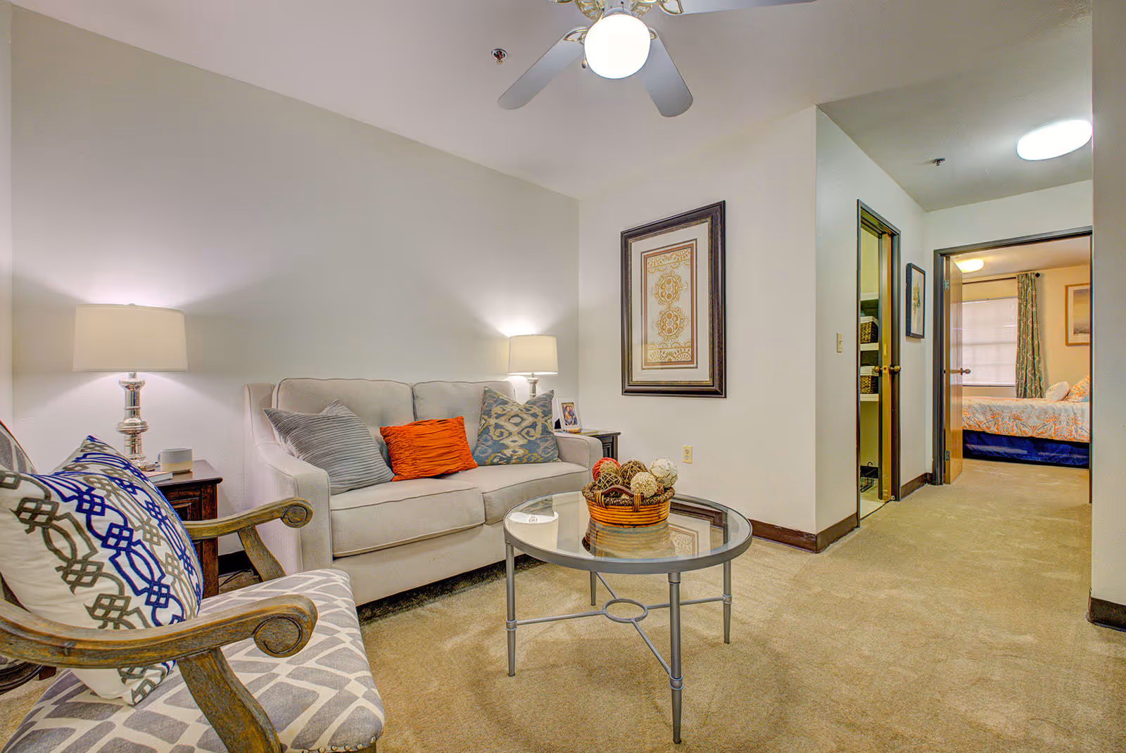 A cozy living room area in an assisted living facility featuring a beige sofa with colorful throw pillows, a wooden armchair with patterned cushions, a round glass coffee table with a decorative basket centerpiece, two table lamps on side tables, a framed artwork on the wall, and a view into a bedroom with a bed and curtains.