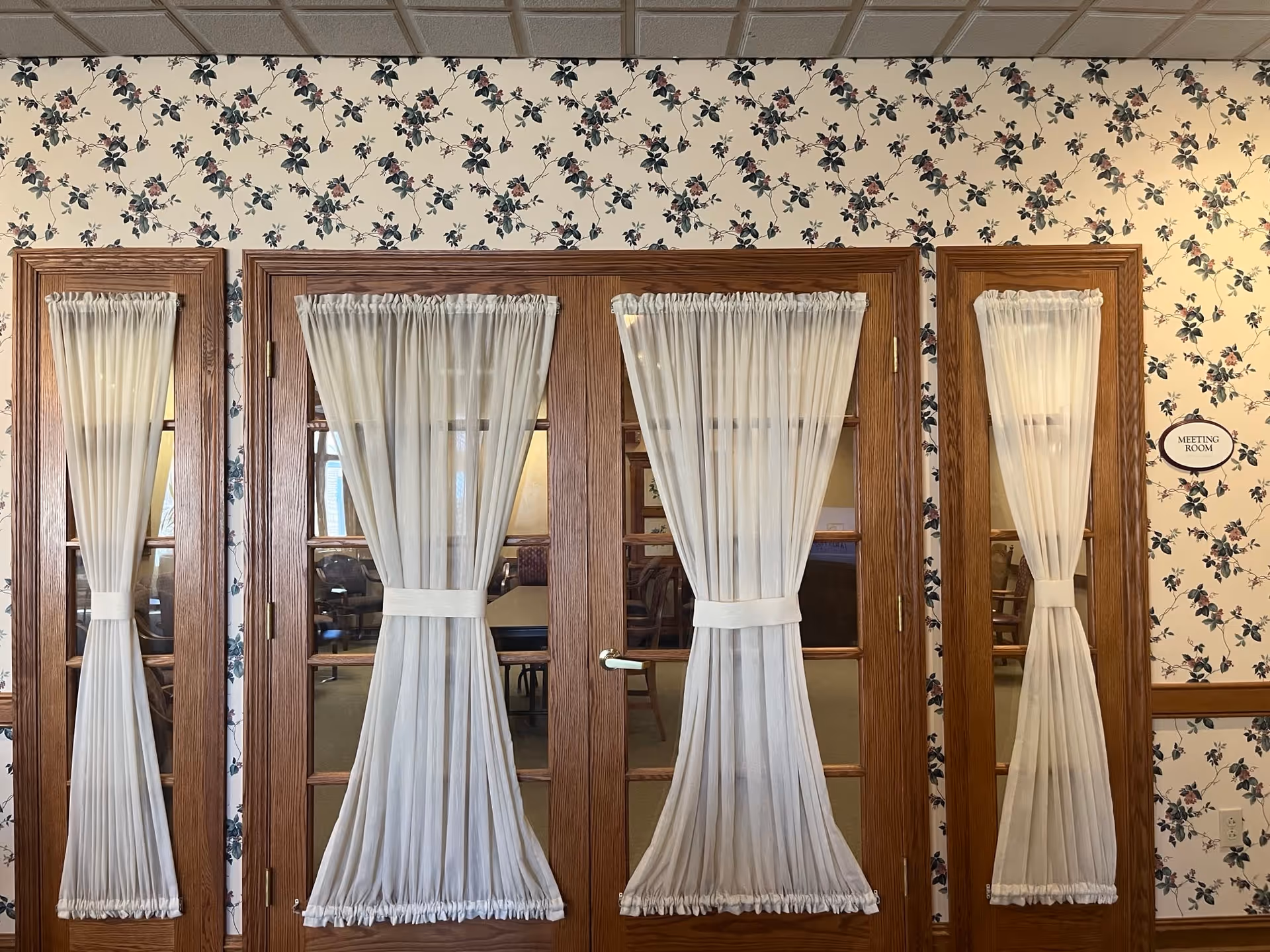 Wooden double doors with glass panels and sheer white curtains tied in the middle, set in a wall with floral wallpaper. A small oval sign to the right of the doors reads 'Meeting Room'.
