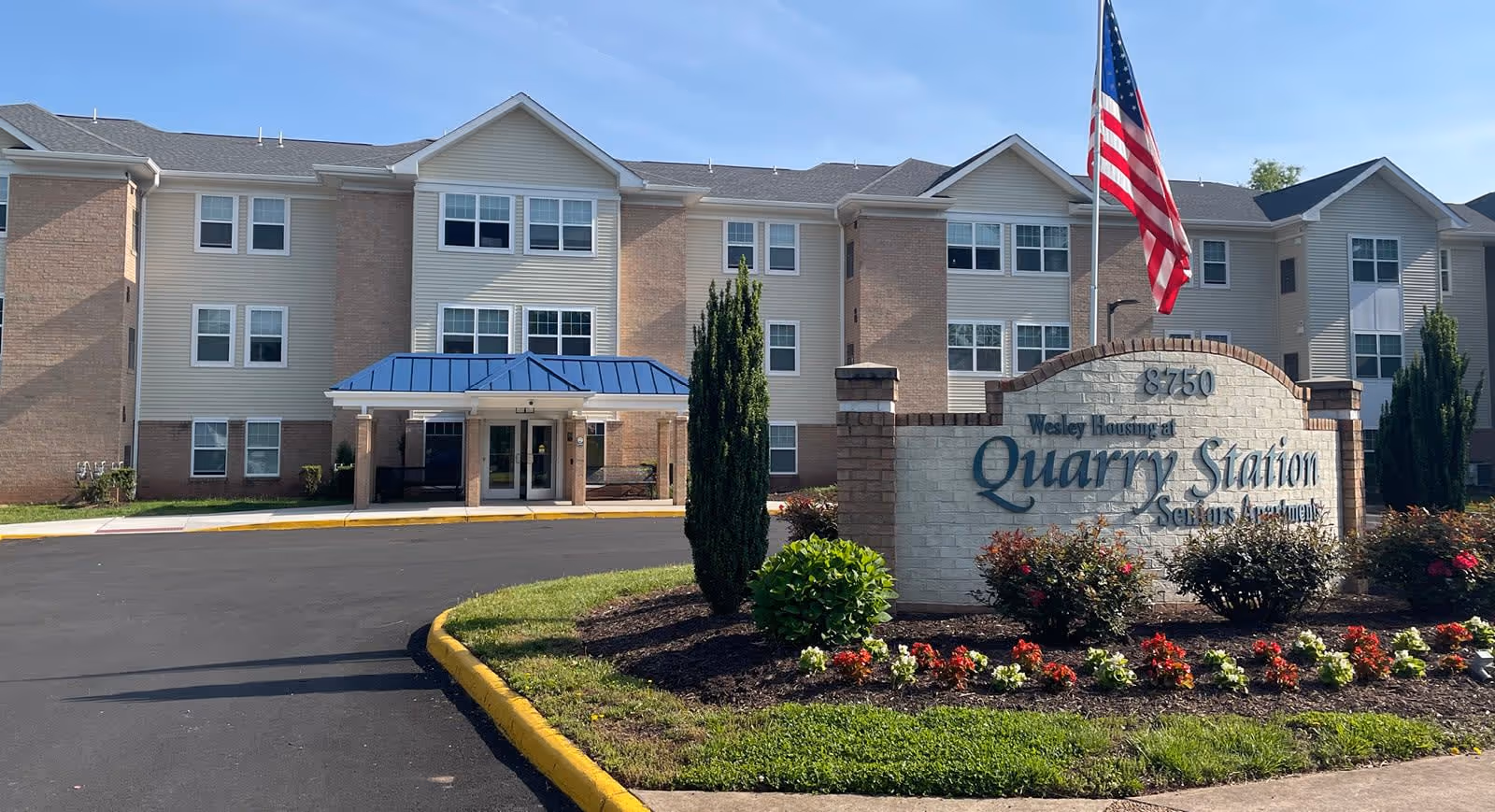 Exterior view of Quarry Station Seniors Apartments building with a three-story structure, beige siding and brick accents, a blue awning over the entrance, and a landscaped area with flowers and bushes in front. An American flag is flying on a pole next to the sign displaying the facility's name and address.