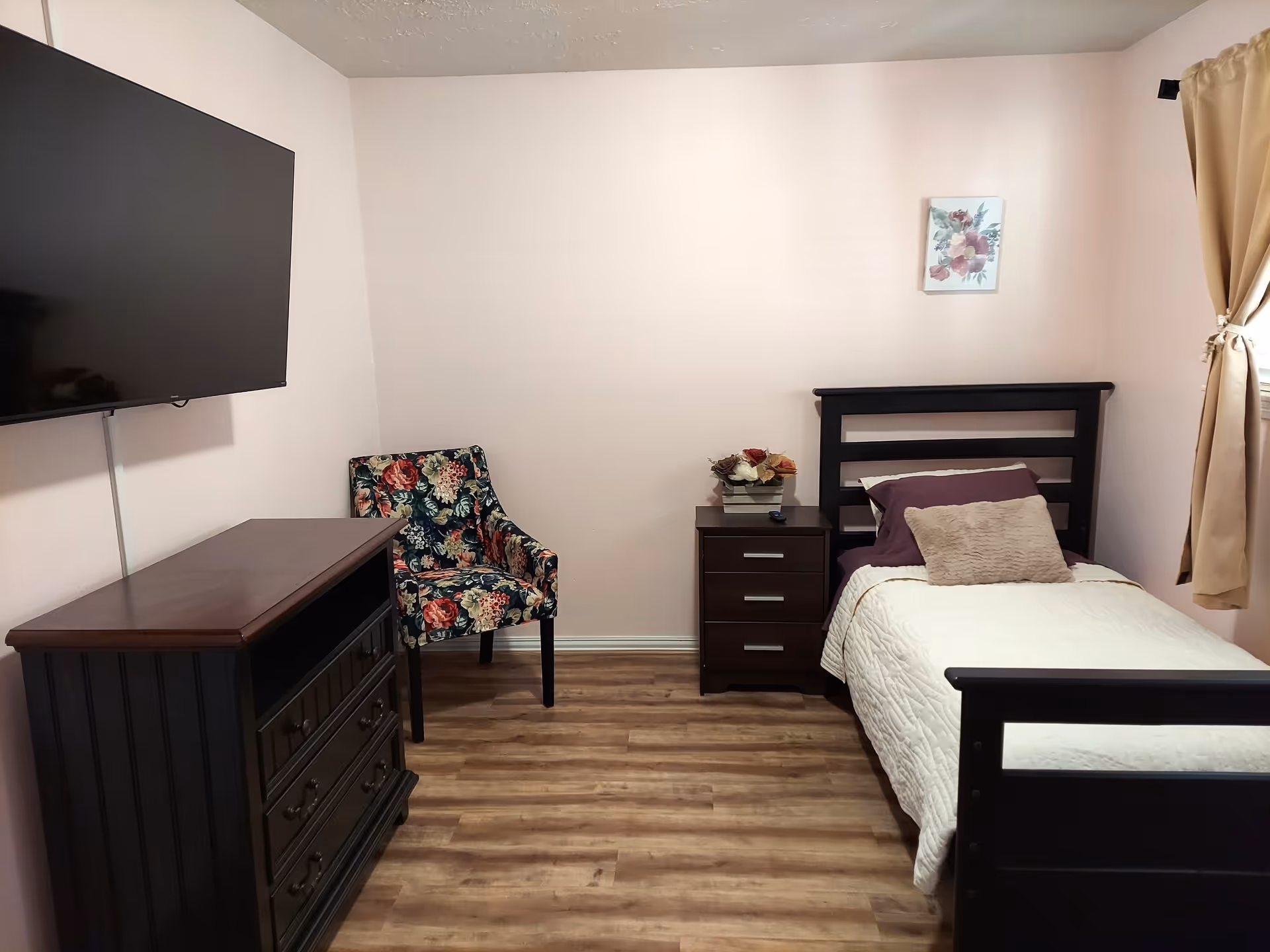 A small bedroom with a single bed featuring dark wooden frame, white quilt, and purple pillows. Next to the bed is a dark wooden nightstand with a flower arrangement on top. A floral-patterned armchair is placed in the corner beside a dark wooden dresser with a flat-screen TV mounted on the wall above it. Light-colored curtains cover a window on the right side of the room, and a small floral painting hangs on the wall above the bed.