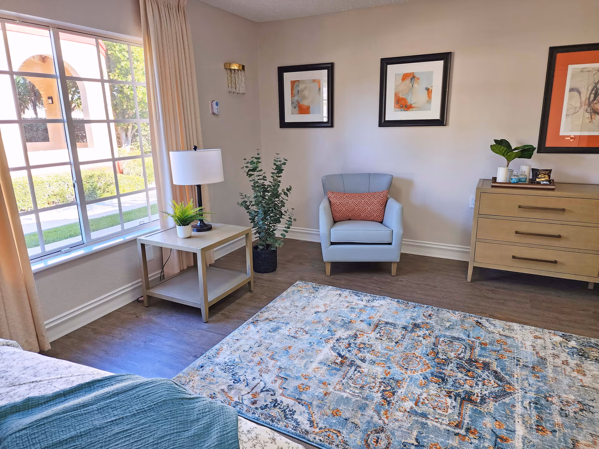A cozy room with a large window letting in natural light, beige curtains, a light blue armchair with an orange patterned pillow, a wooden side table with a lamp and a small plant, a potted plant on the floor, a wooden dresser with decorative items, and a large patterned area rug on a wooden floor. Three framed abstract artworks hang on the wall.