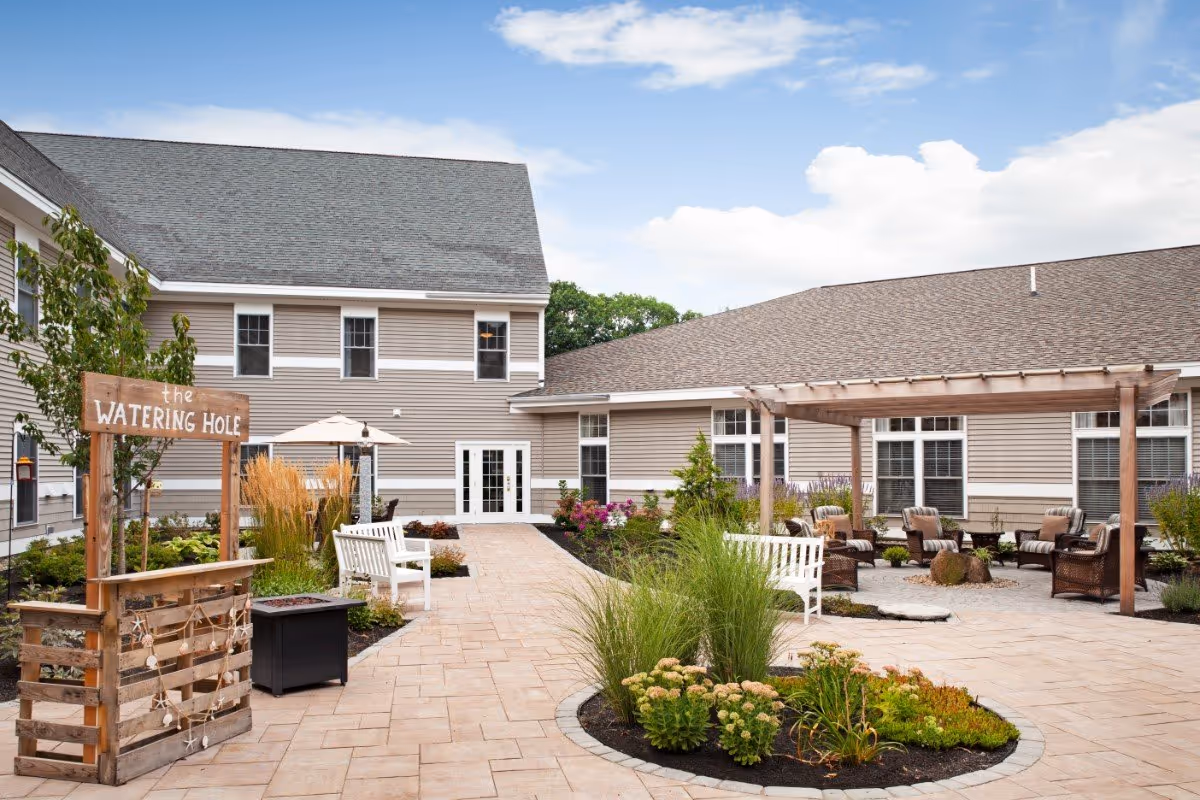 Outdoor courtyard area of a senior living facility with paved walkways, garden beds with various plants, white benches, a wooden pergola with cushioned chairs underneath, and a wooden sign that reads 'the WATERING HOLE'. The building surrounding the courtyard has beige siding and multiple windows under a partly cloudy sky.