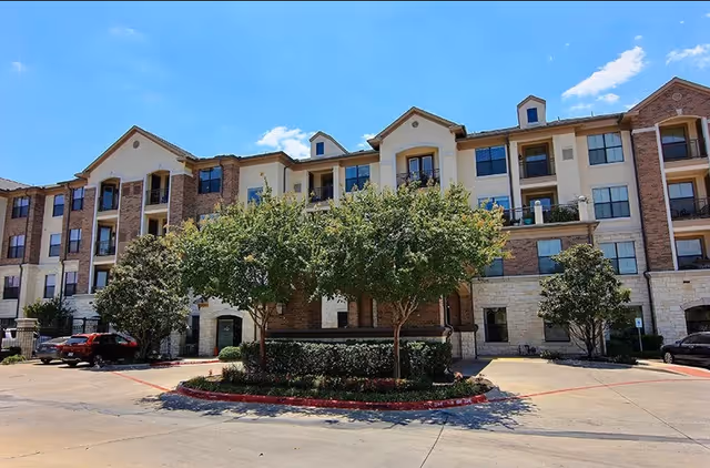 Exterior view of a multi-story senior living facility building with brick and light-colored walls, several windows, balconies, and a small landscaped area with trees and shrubs in front. Cars are parked along the driveway under a clear blue sky.
