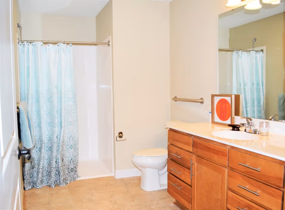 Bright bathroom with a wooden vanity and sink, toilet, and a shower with a blue patterned curtain.
