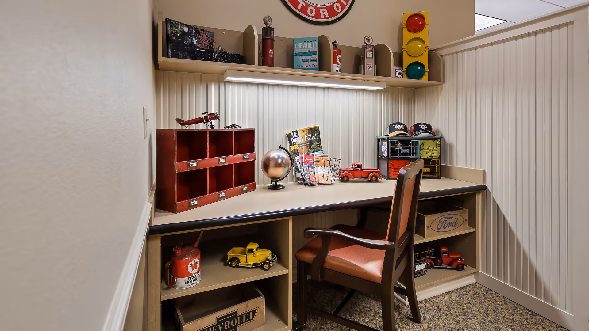 A small office or study nook with a wooden desk and chair. The desk has vintage-style decorations including toy trucks, a globe, a red cubby organizer, a wire basket with books, and a shelf above holding retro gas pumps and a traffic light. The walls have white beadboard paneling and the floor is carpeted.
