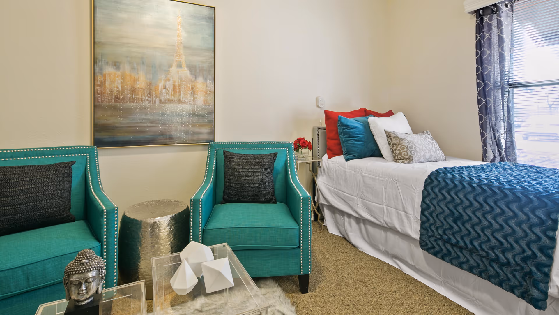 A cozy bedroom featuring a single bed with white bedding, colorful pillows in red, teal, and patterned designs, and a teal textured throw blanket. Next to the bed are two teal armchairs with black cushions, separated by a small metallic side table. A decorative Buddha head sculpture and geometric white ornaments are on a clear table in the foreground. A large window with patterned curtains allows natural light into the room, and a framed artwork hangs on the wall behind the chairs.