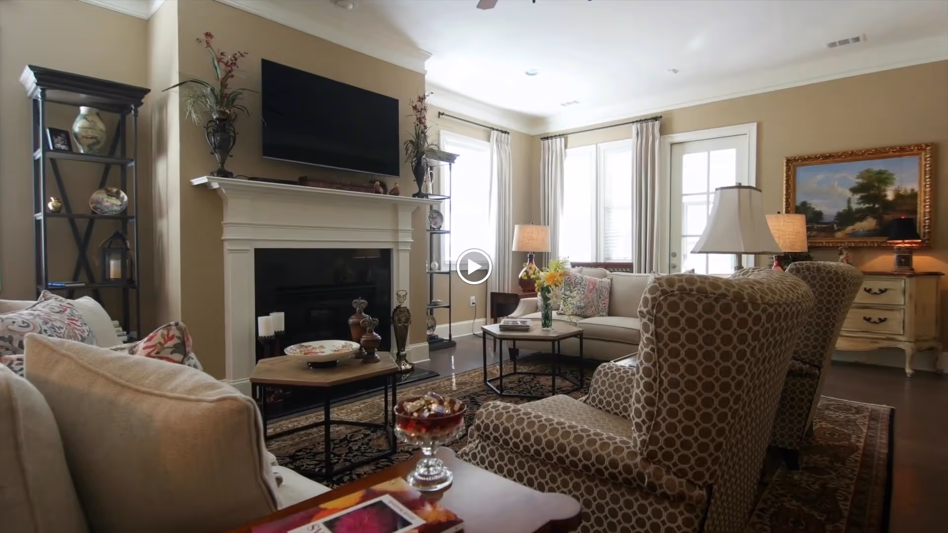 A cozy living room with beige walls and dark hardwood floors. The room features a white fireplace with a flat-screen TV mounted above it, flanked by two black shelving units holding decorative items. There are two patterned armchairs and a beige sofa with floral cushions arranged around a coffee table. The room is well-lit with natural light coming through windows with white curtains, and there are two table lamps providing additional lighting. A framed landscape painting hangs on the wall above a small chest of drawers.
