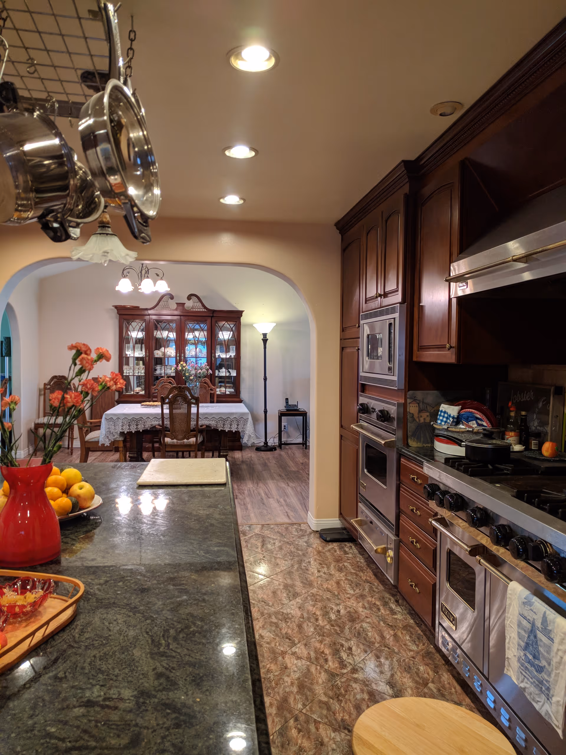 Spacious kitchen with a dark granite island, hanging pots, stainless steel range and wooden cabinets opening to a dining room with a china cabinet.