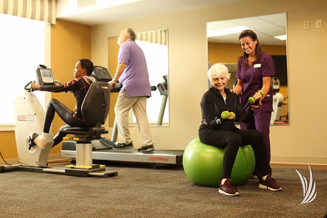 A senior woman exercises by lifting green dumbbells while sitting on a green exercise ball, assisted by a smiling caregiver in purple scrubs. In the background, another senior woman uses a recumbent exercise bike and a senior man walks on a treadmill in a well-lit fitness room with mirrors on the walls.