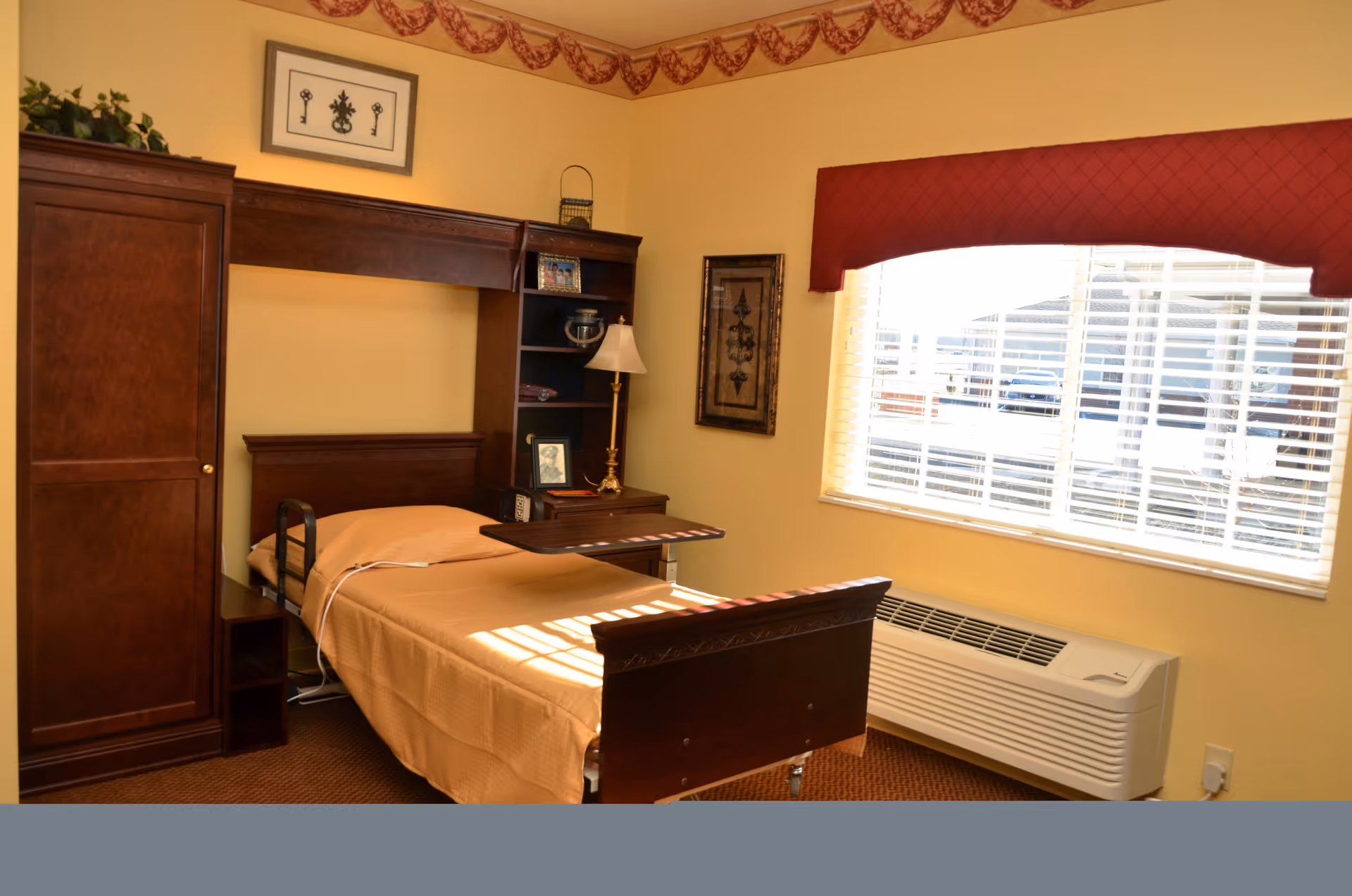 Sunlit senior living bedroom with a single bed, wooden headboard and shelving unit, bedside lamp, and a window with blinds and an air conditioner.