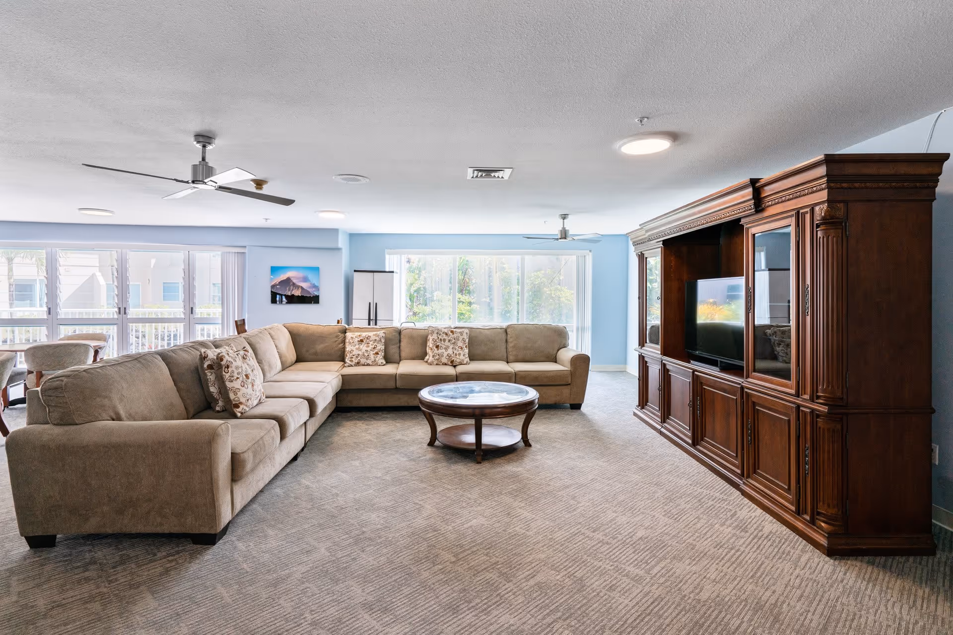 A spacious living room with a large beige sectional sofa adorned with patterned cushions, a round wooden coffee table with a glass top, and a large wooden entertainment center housing a flat-screen TV. The room has light blue walls, large windows letting in natural light, ceiling fans, and carpeted flooring.
