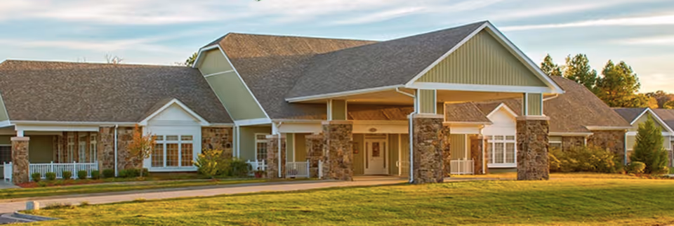 Exterior view of a single-story assisted living facility building with a covered entrance supported by stone pillars, surrounded by a well-maintained lawn and some shrubs under a partly cloudy sky.