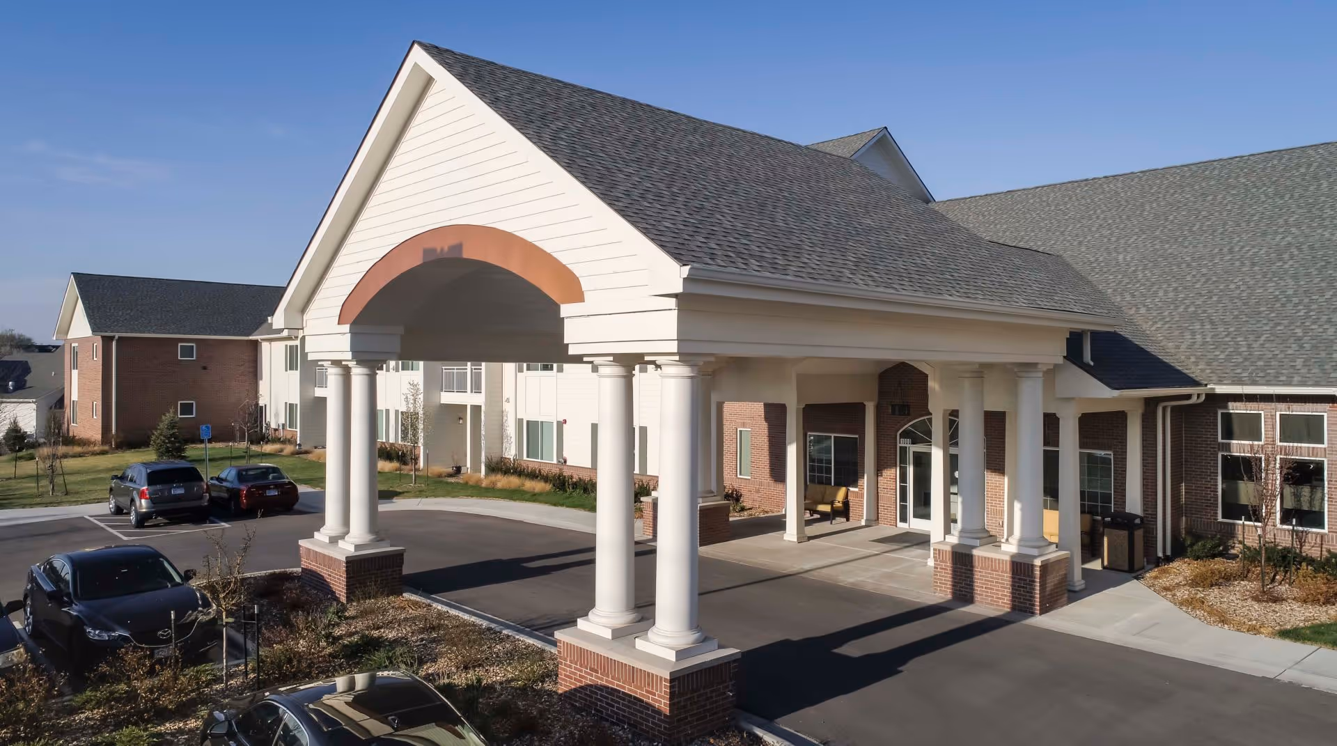 Exterior view of Pioneer Ridge Independent Living of Lawrence showing the main entrance with a covered driveway supported by white columns, brick and siding building facade, parked cars, and a clear blue sky.