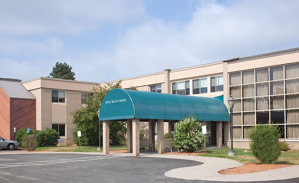 Front exterior of a low-rise brick care facility with a green arched entrance canopy reading "APPLE VALLEY CENTER" and an adjacent parking area.