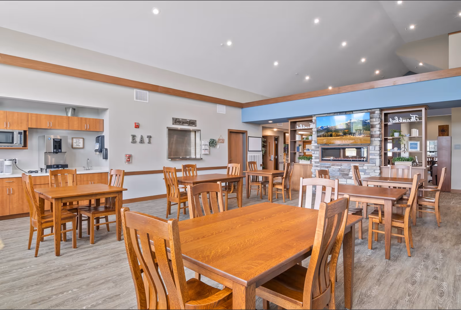 A spacious dining room with multiple wooden tables and chairs arranged neatly. The room features a kitchenette area with cabinets, a microwave, and a coffee machine. On the wall, there are decorative letters spelling 'EAT' and a sign that says 'Come special home.' A stone fireplace with a mounted flat-screen TV is visible in the background, along with shelves holding plants and decorative items. The ceiling is high with recessed lighting, and the floor has a wood-like finish.