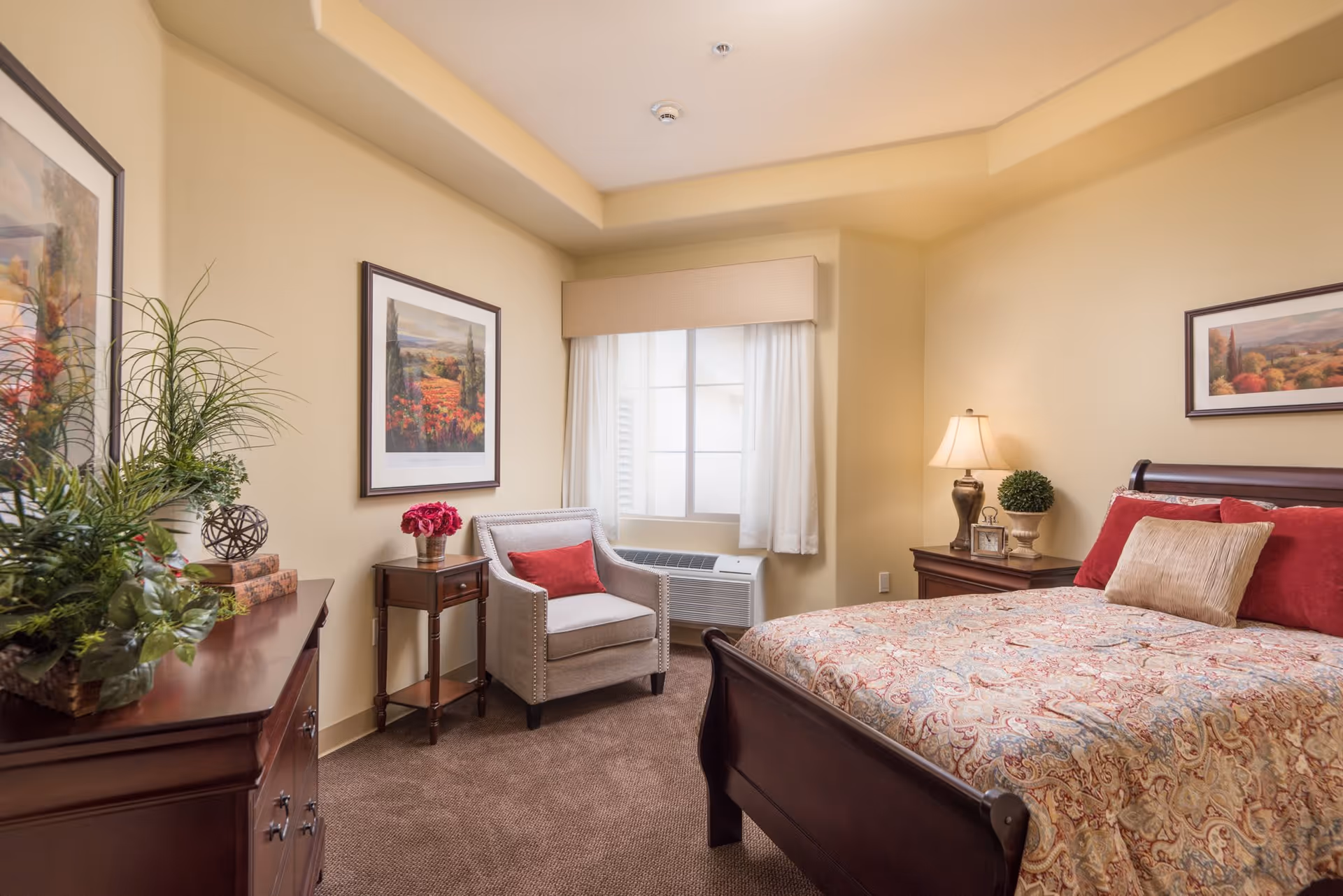 A cozy bedroom with a large wooden bed covered in a patterned bedspread and red and beige pillows. There is a wooden nightstand with a lamp, a small plant, and a clock. A beige armchair with a red pillow sits next to a window with white curtains. On the left side, there is a wooden dresser with decorative plants and framed artwork on the walls.