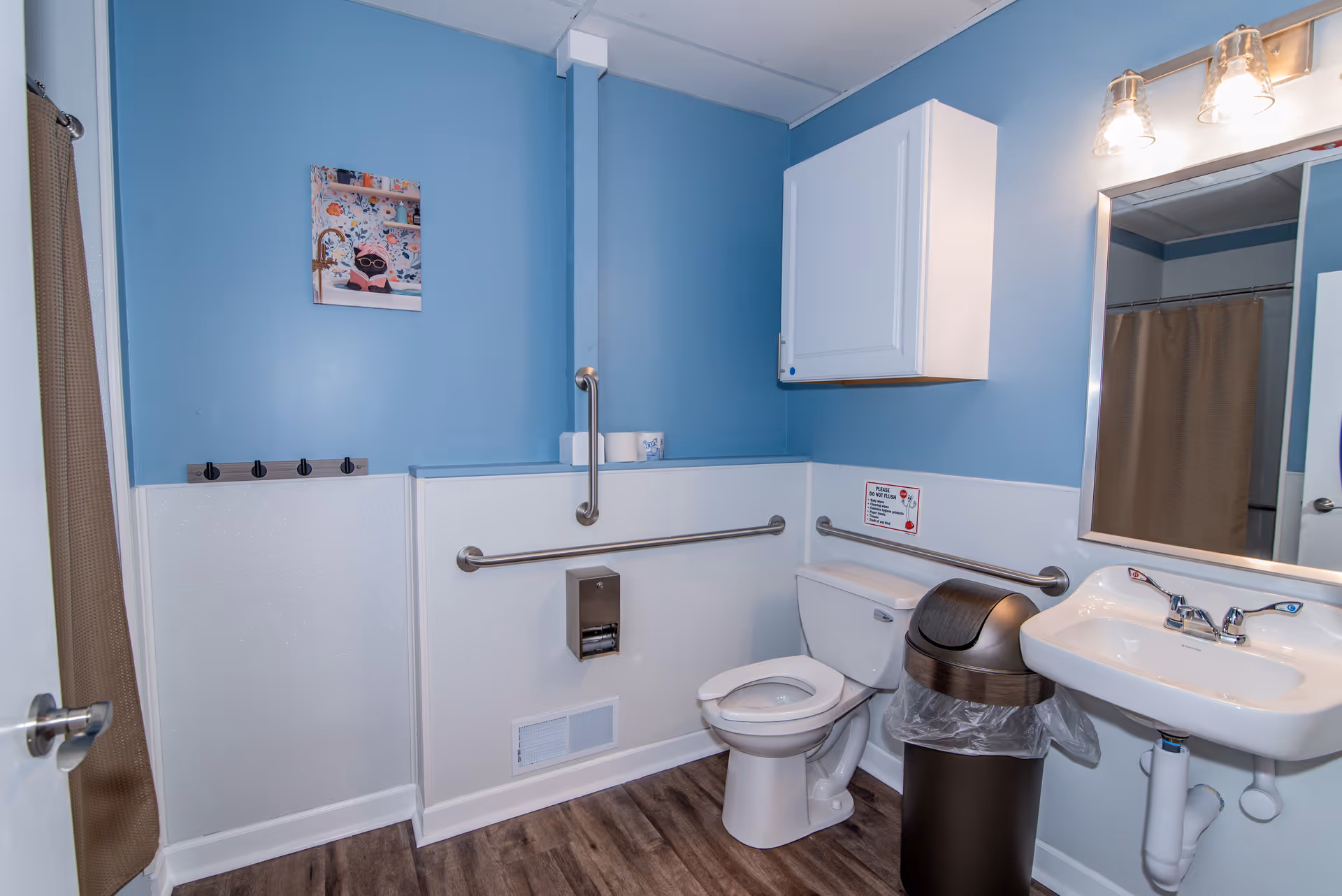 A clean and accessible bathroom with blue walls and wood-look flooring. The bathroom features a white toilet with grab bars on the walls, a white sink with a mirror above it, a trash can, a white cabinet mounted on the wall, and a shower with a brown curtain. There is a small colorful picture on the wall and a sign above the toilet with flushing instructions.