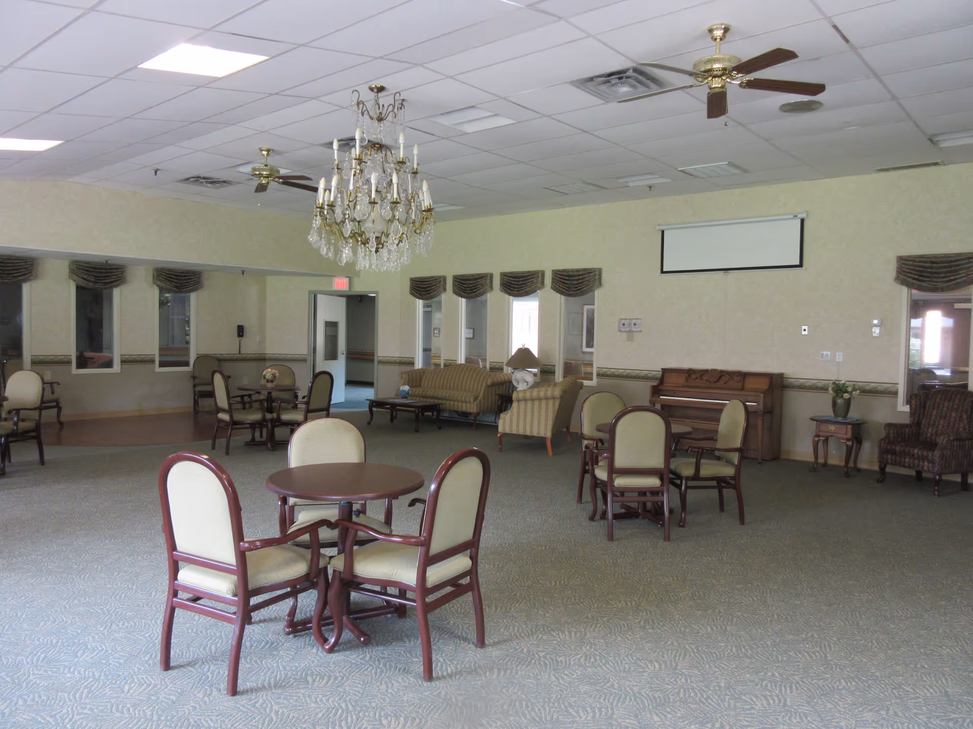 A spacious common area with multiple round tables and chairs, a crystal chandelier hanging from the ceiling, ceiling fans, a piano against the wall, and sofas with a lamp. The room has carpeted flooring and windows with valances.