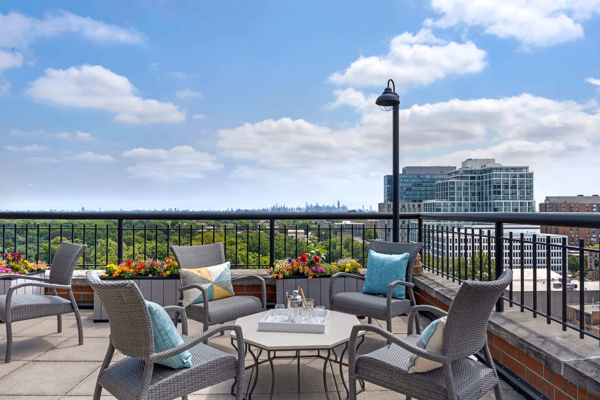 Rooftop terrace with wicker chairs and a table, flower planters, and a distant city skyline under a blue sky.