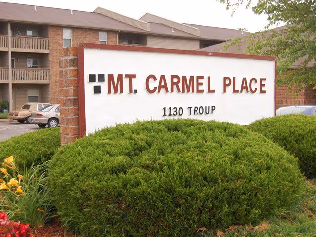 Outdoor view of a sign for Mt. Carmel Place located at 1130 Troup, surrounded by green bushes and flowers with a multi-story residential building and parked cars in the background.