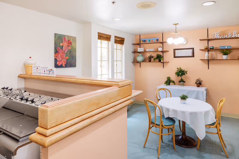A cozy dining area with a round table and chairs, decorated with plants and shelves in a senior living facility.