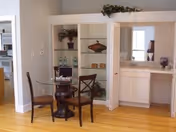 A small dining area with a round glass table and two wooden chairs on a hardwood floor. Behind the table is a white built-in cabinet with open shelves displaying decorative items and bottles. To the right, there is a vanity area with a mirror, countertop, and cabinets below. The room has light-colored walls and a doorway leading to another room.