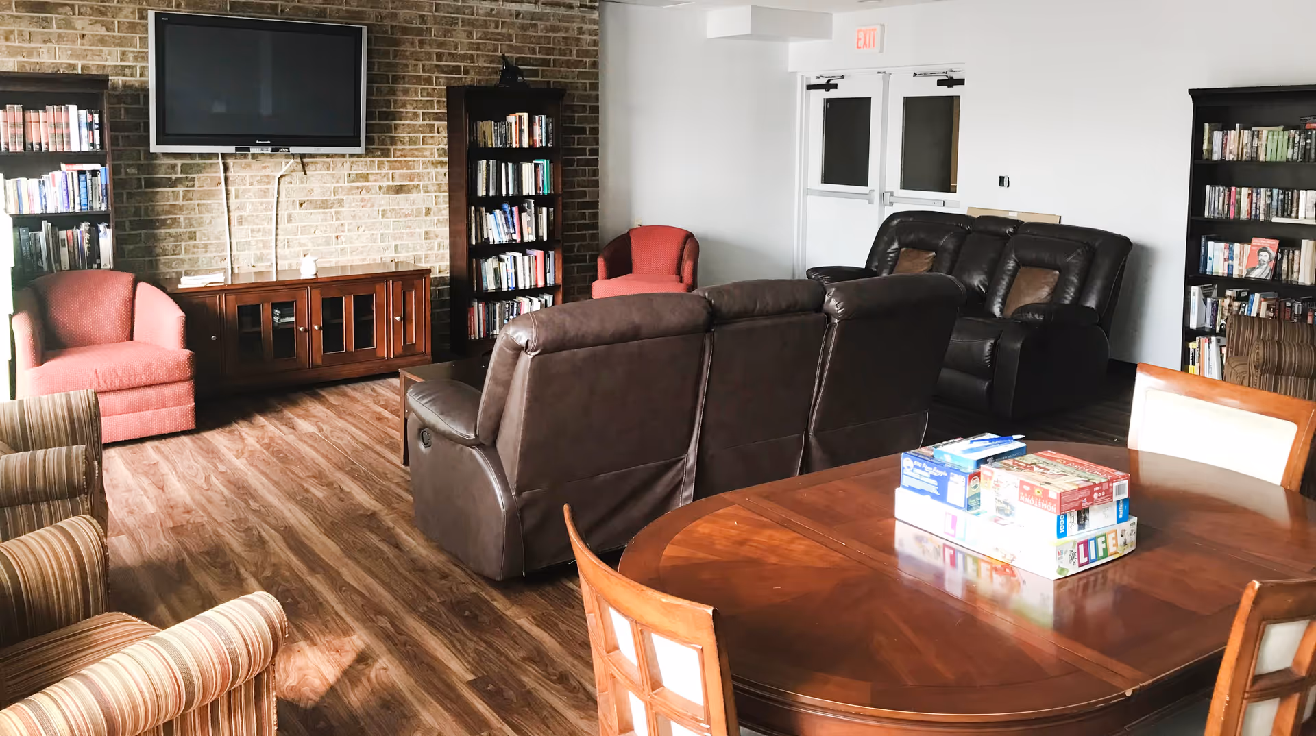 A cozy common living area with brown leather recliners, red and striped armchairs, wooden bookshelves filled with books, a wall-mounted flat screen TV, and a wooden table with board games on top. The room has wood flooring and white walls with a brick accent wall behind the TV.