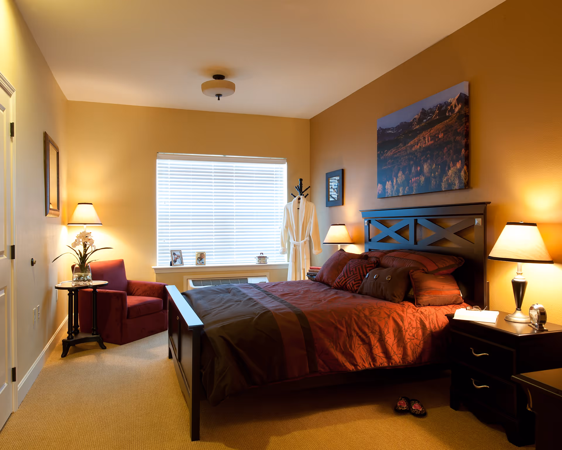 A cozy bedroom with warm yellow walls, a large window with blinds, a dark wooden bed with red and brown bedding, two bedside tables with lamps, a red armchair next to a small round table with a vase of flowers, a white robe hanging on a coat rack, and framed pictures on the window sill.